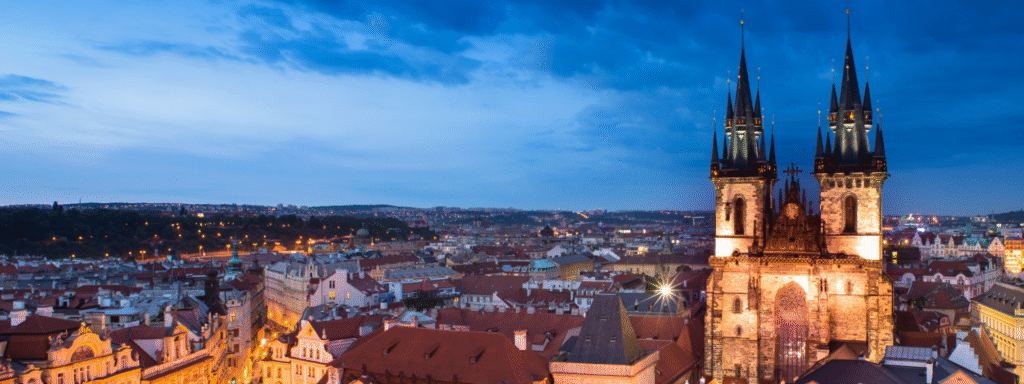 Cityscape at dusk featuring a large cathedral with two tall spires overlooking red-roofed buildings and illuminated streets.