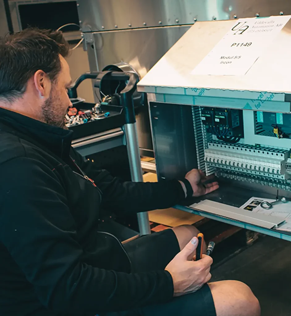Person seated beside an open electrical cabinet, holding tools and inspecting components inside a metal enclosure.