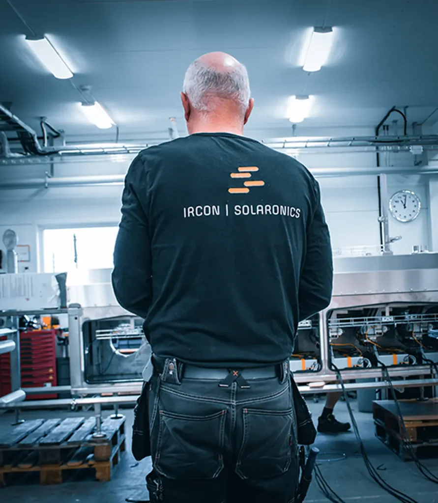 Person standing in a workshop wearing a black shirt with the Ircon Solaronics logo, facing industrial equipment.