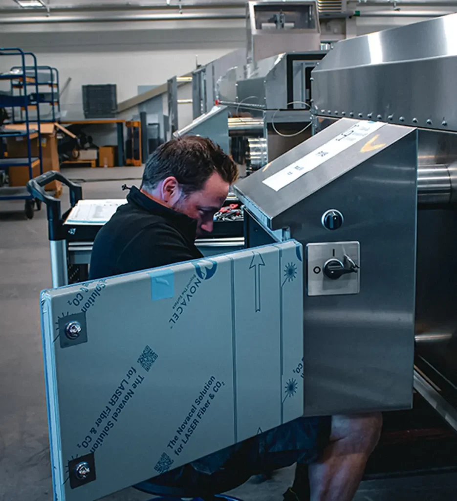 Person seated on the floor working on a large metallic machine with an open panel in an industrial workspace.